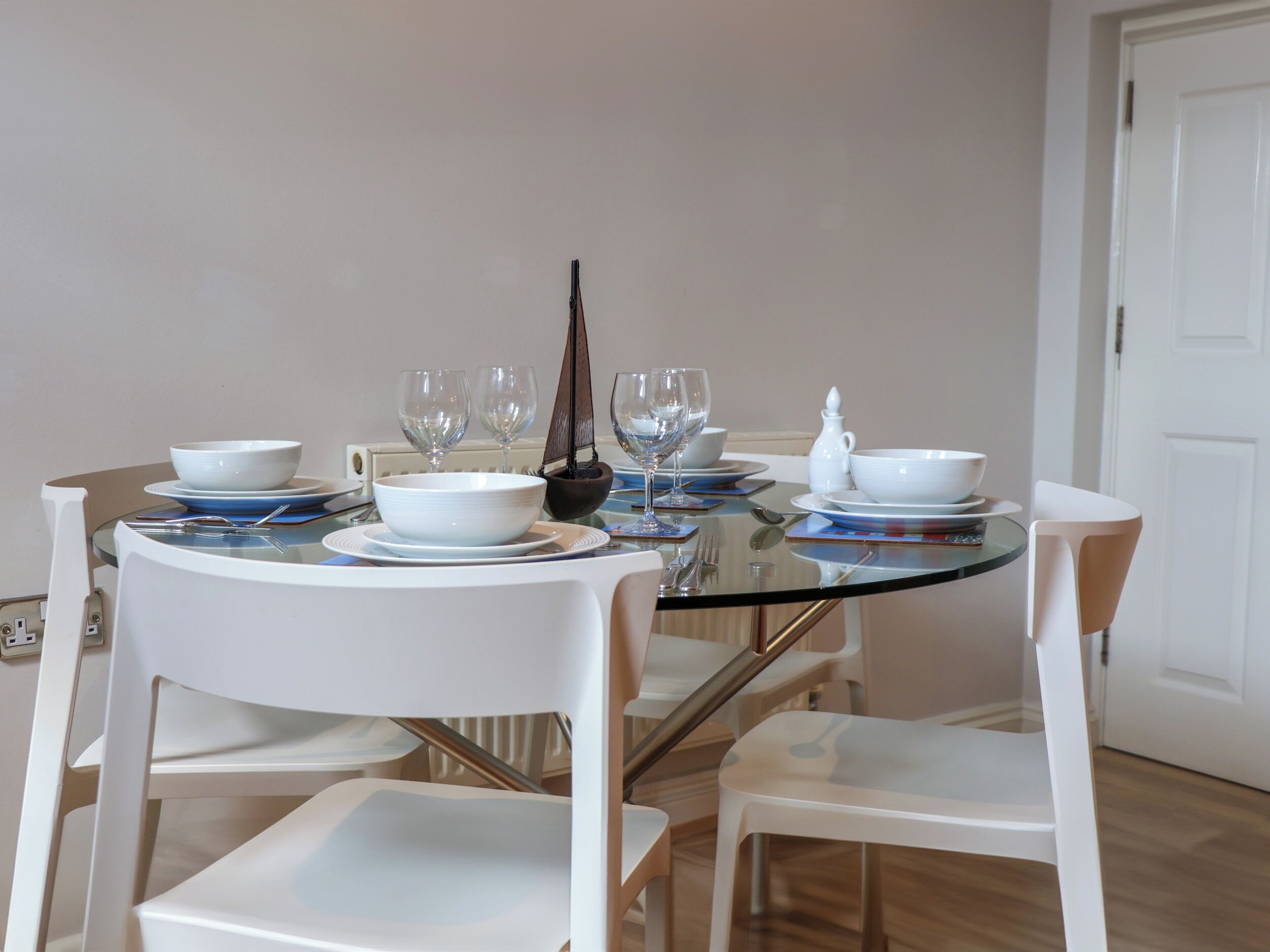 A dining table with white chairs, clear glass top, and white plates.