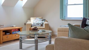 Una sala de estar acogedora con techo alto, una mesa de centro de vidrio, un sofá color beige y una ventana con contraventanas.
