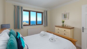 A bedroom with a large bed, a dresser, a mirror, and a window with a view of the sea. - Cornerways (St Ives)