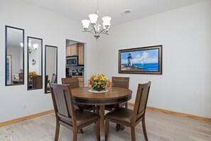 A dining area with a round wooden table, six chairs, and a chandelier. There is a painting of a lighthouse on the wall.