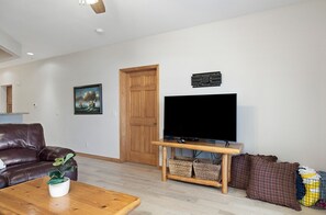A living room with a brown leather sofa, a wooden coffee table with a plant, a TV on a wooden stand, a wooden door, and a painting on the wall.
