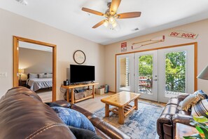 A living room with a leather sofa, a wooden coffee table, a flat-screen TV, and a ceiling fan. There is a sliding glass door leading to an outdoor area with a patio table and chairs.
