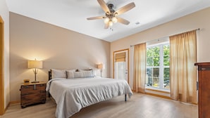 Ein Schlafzimmer mit Bett, Deckenventilator, Nachttisch, Lampe und einem Fenster mit Vorhängen.