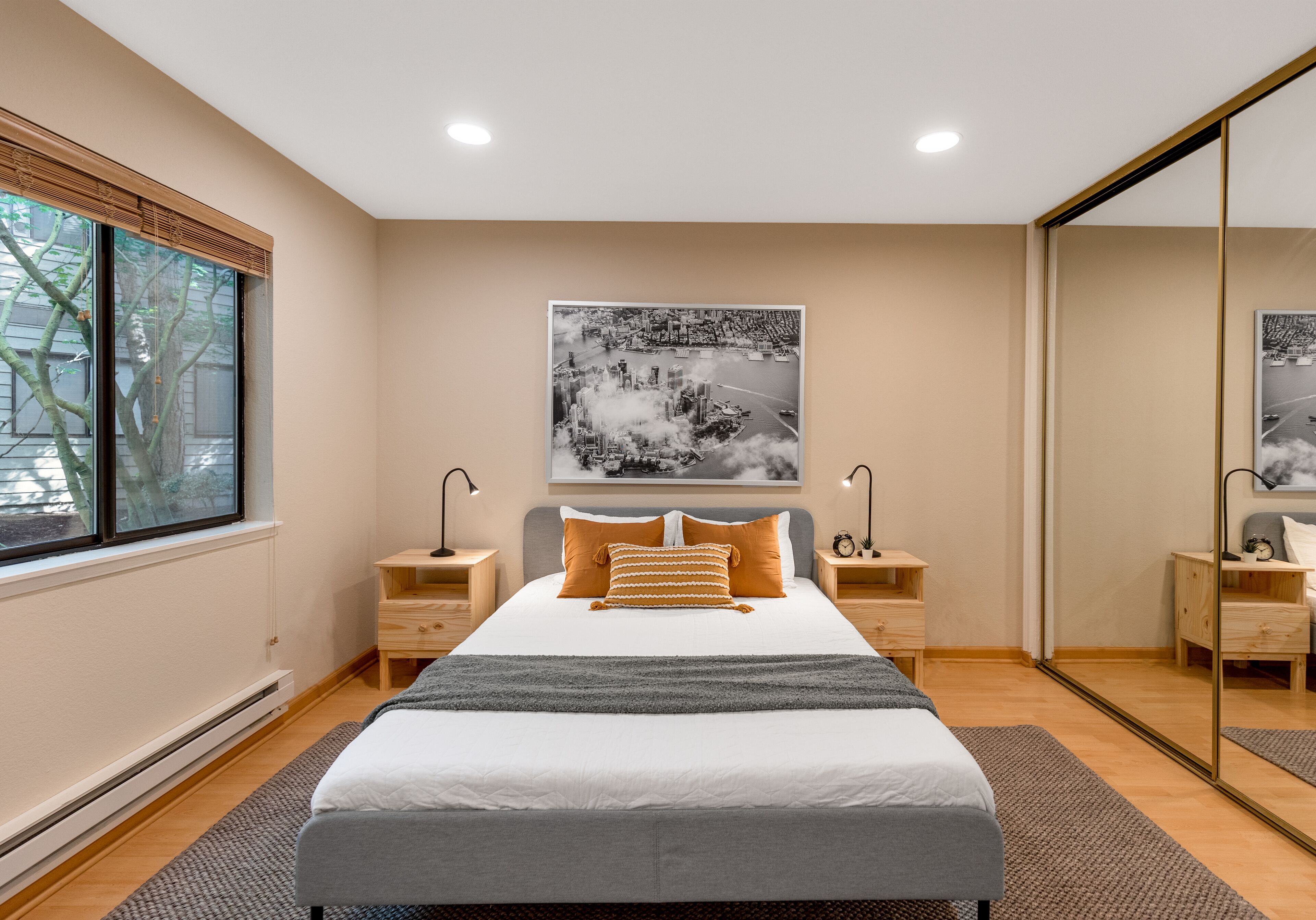 A modern bedroom with a large bed, two bedside tables, a mirror, and a cityscape painting on the wall.