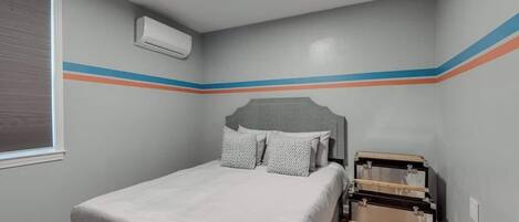 Une chambre à coucher avec un lit, une fenêtre avec des stores, un climatiseur et une commode en bois.