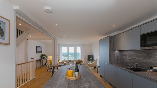 A modern kitchen with a central island, a dining area with a view, and a staircase leading to a balcony. - Beach View (St Ives)