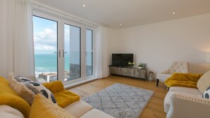 A spacious living room with a large sliding glass door leading to a balcony with a view of the sea. - Beach View (St Ives)