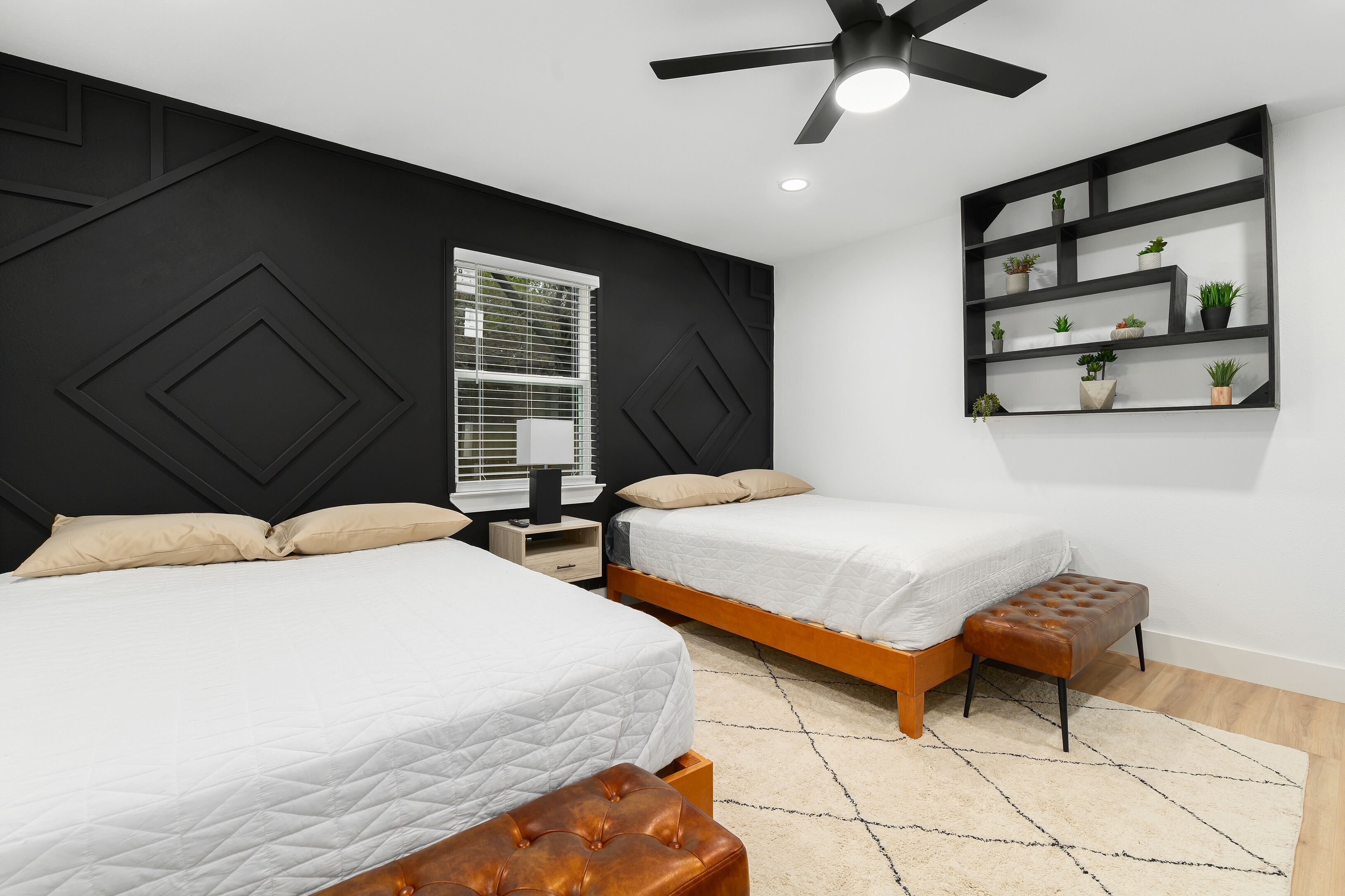 A modern bedroom with two beds, a ceiling fan, a window with blinds, a bedside table, and a wall-mounted plant shelf.