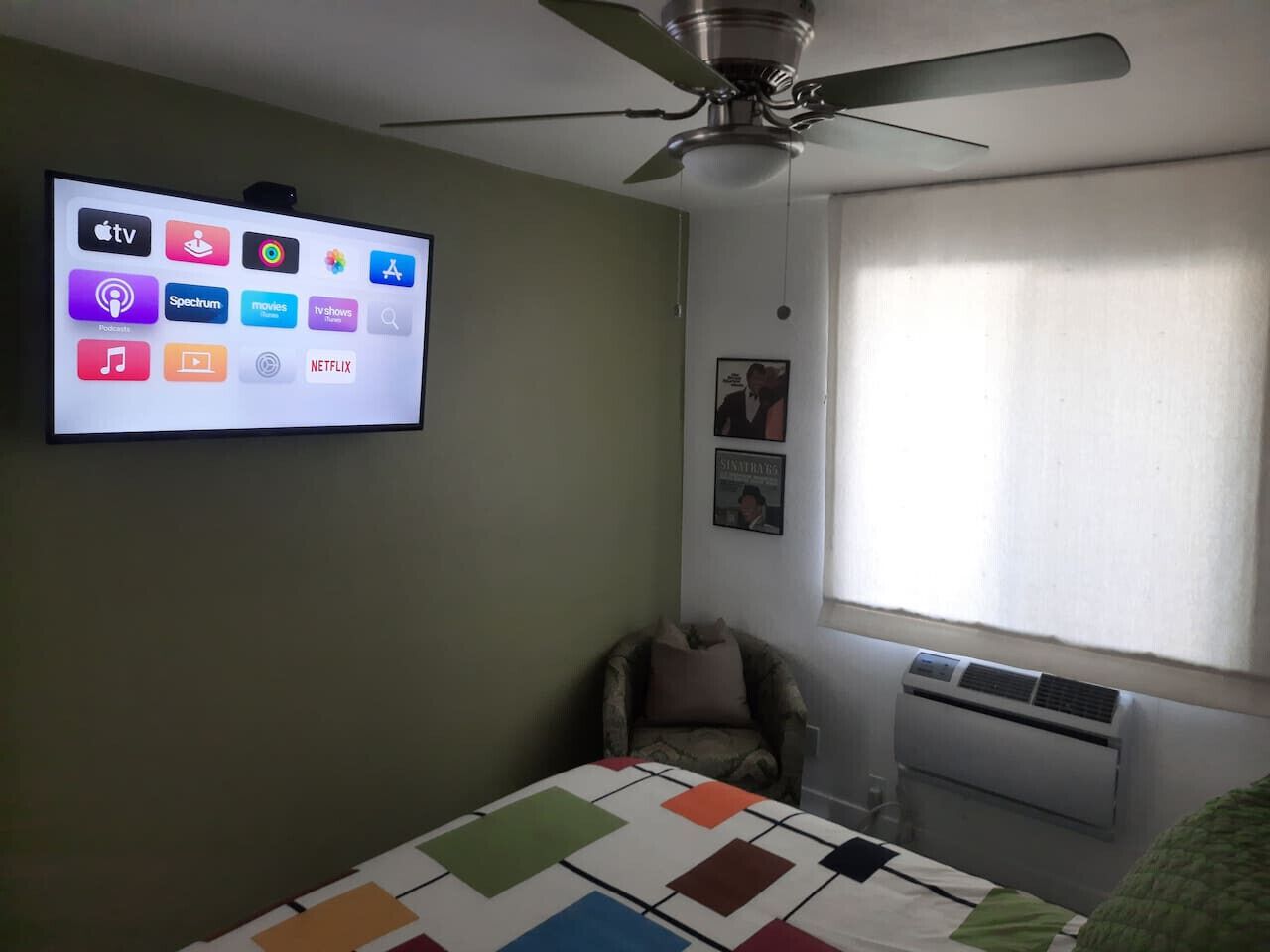 A bedroom with a bed, a chair, a ceiling fan, an air conditioner, and a TV displaying streaming services.