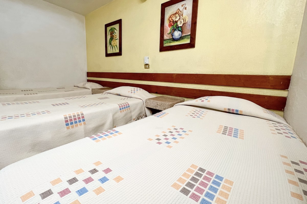 A hotel room with two single beds, a wooden headboard, a wall-mounted light, and two framed floral paintings.