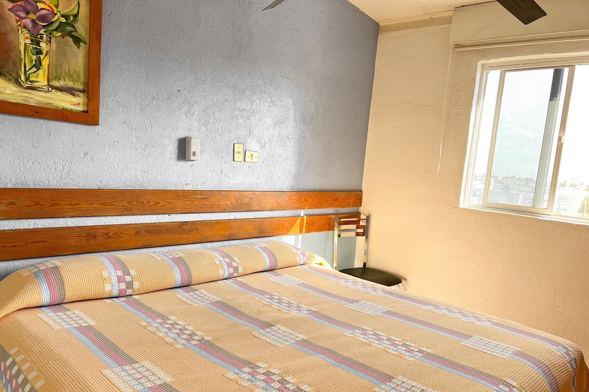 A neatly made bed with a patterned bedspread, a wooden headboard, a wall-mounted painting, and a window with a view.