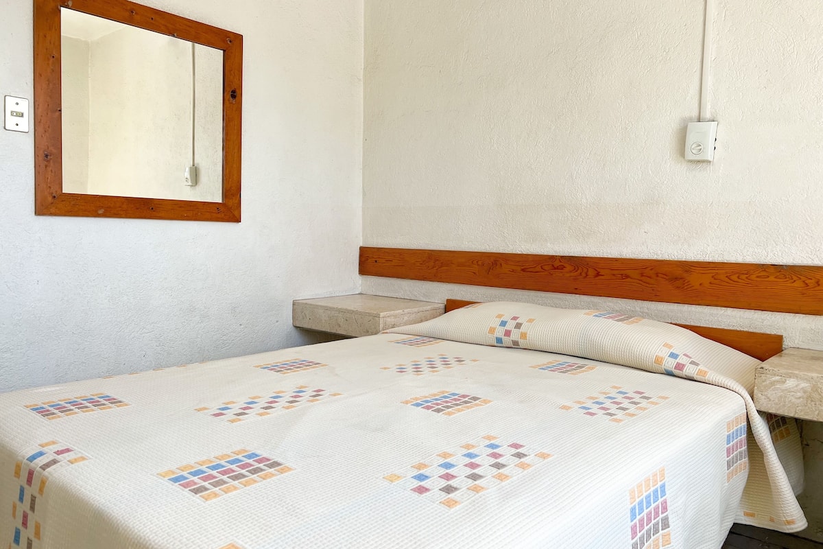 A bedroom with a wooden headboard, a bedside table, a nightstand, and a bed with a quilt featuring a geometric pattern.