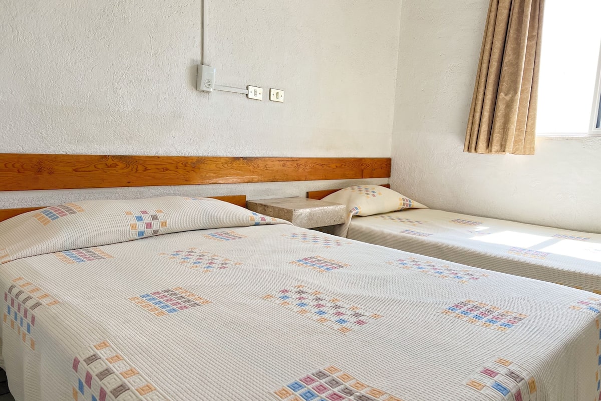 A room with two beds, a wooden headboard, a window with curtains, and a wall-mounted electrical outlet.