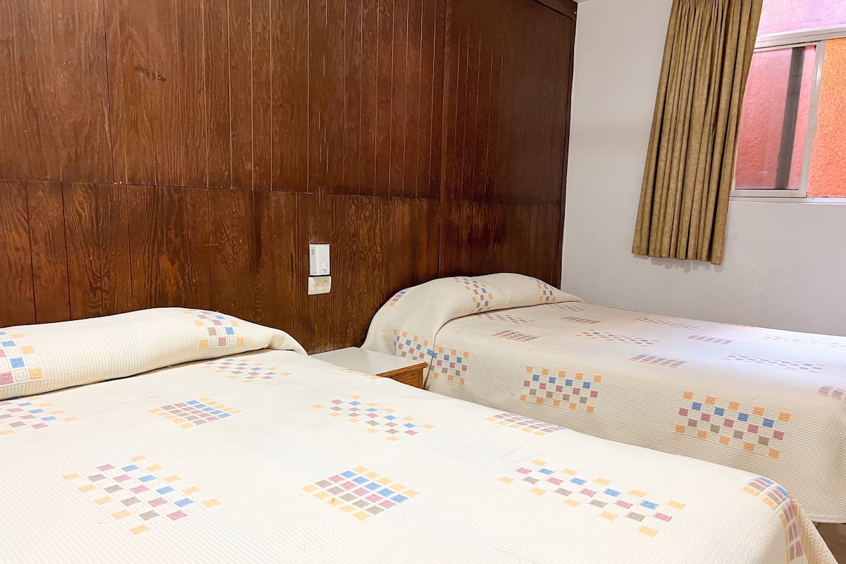 A room with two beds, wooden headboard, a window with curtains, and a thermostat on the wall.