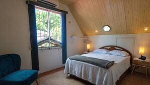 Ein Schlafzimmer mit schrägen Wänden, einem Bett mit weißen Bettwäsche, einem Nachttisch mit Lampe, einem Sessel und einem Fenster mit Blick auf Grünflächen.