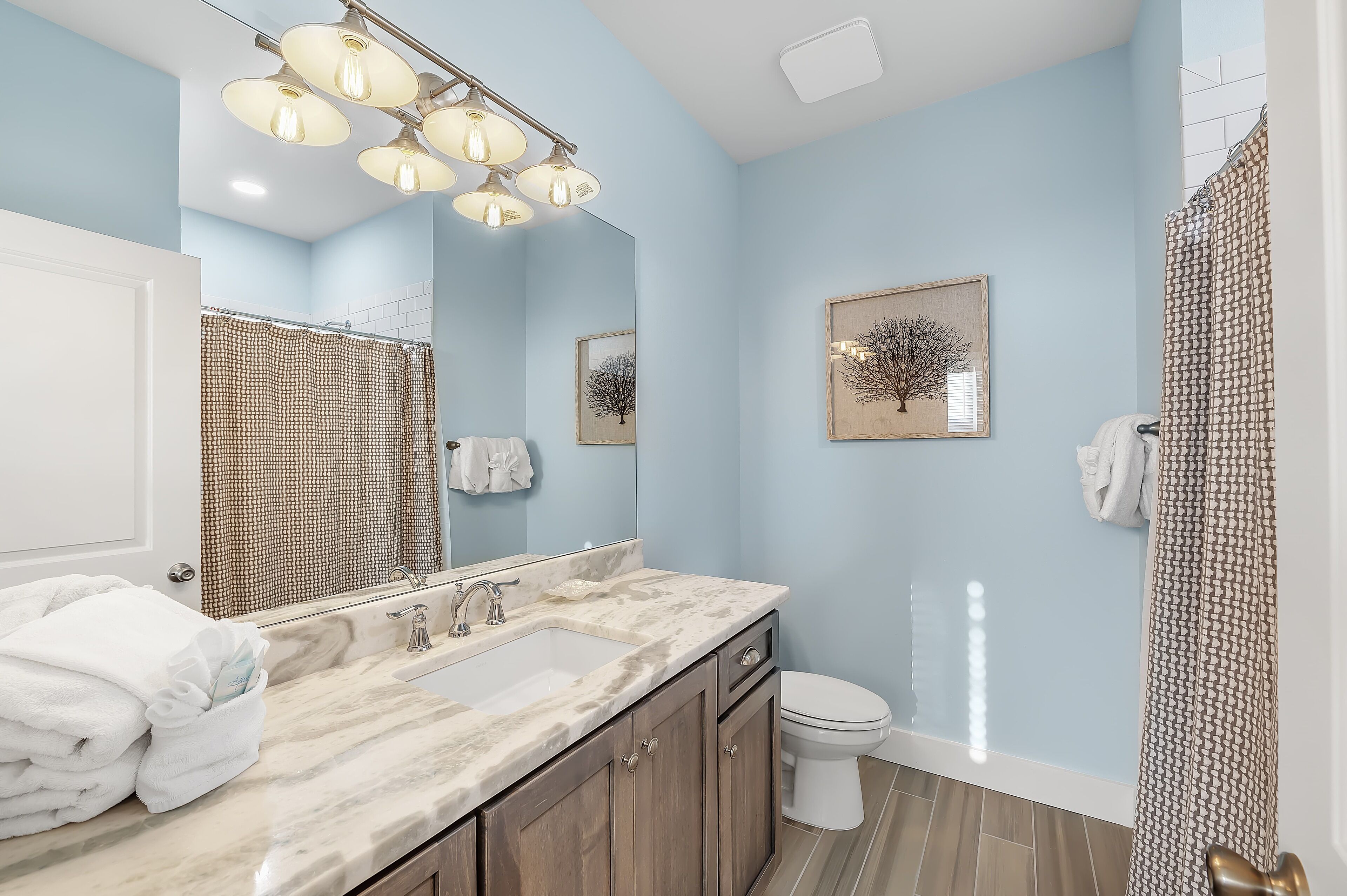 Une salle de bain avec un lavabo en marbre, un grand miroir, une douche avec un rideau à motifs et deux tableaux encadrés au mur.