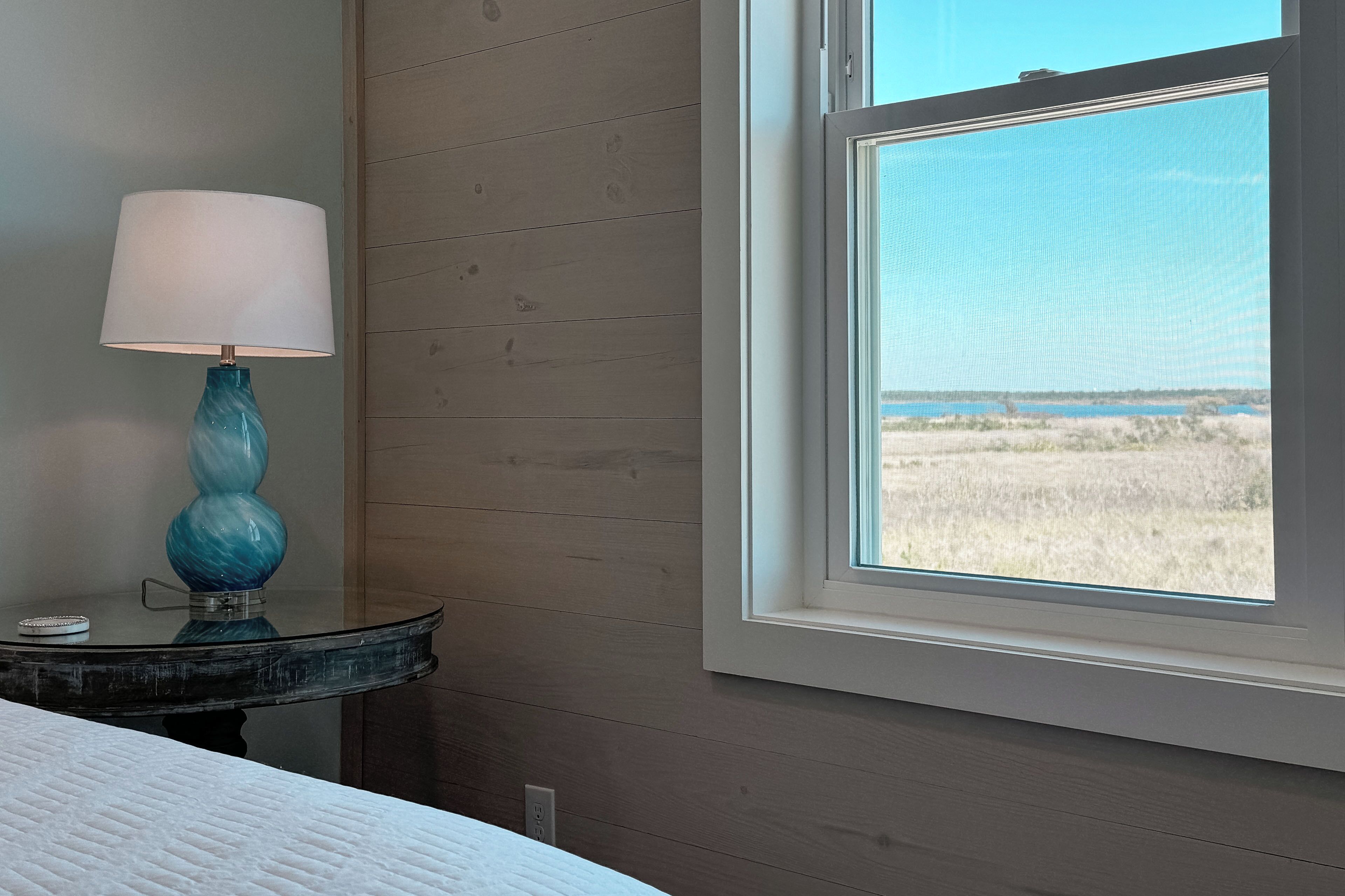 A room with a bed, a lamp on a table, and a window with a view of a beach.