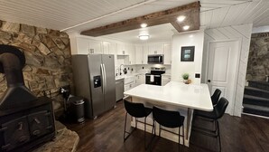 A modern kitchen with white cabinets, a central island, black chairs, a stone wall, and a built-in wood stove. - The Cottage in the Cove (Denver)