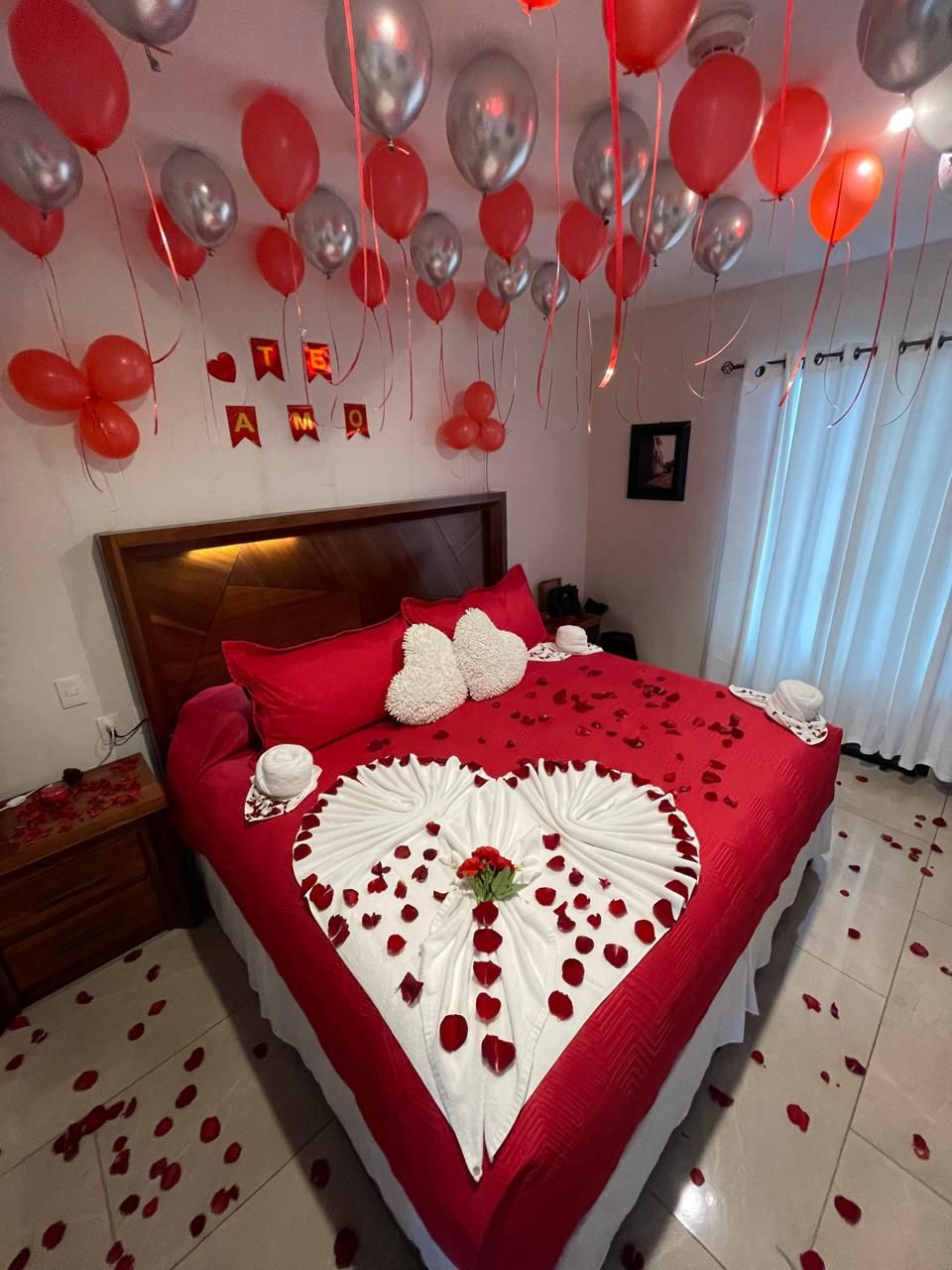 Una cama con una colcha roja, almohadas blancas y un arreglo de pétalos de rosa en forma de corazón y un ramo.
