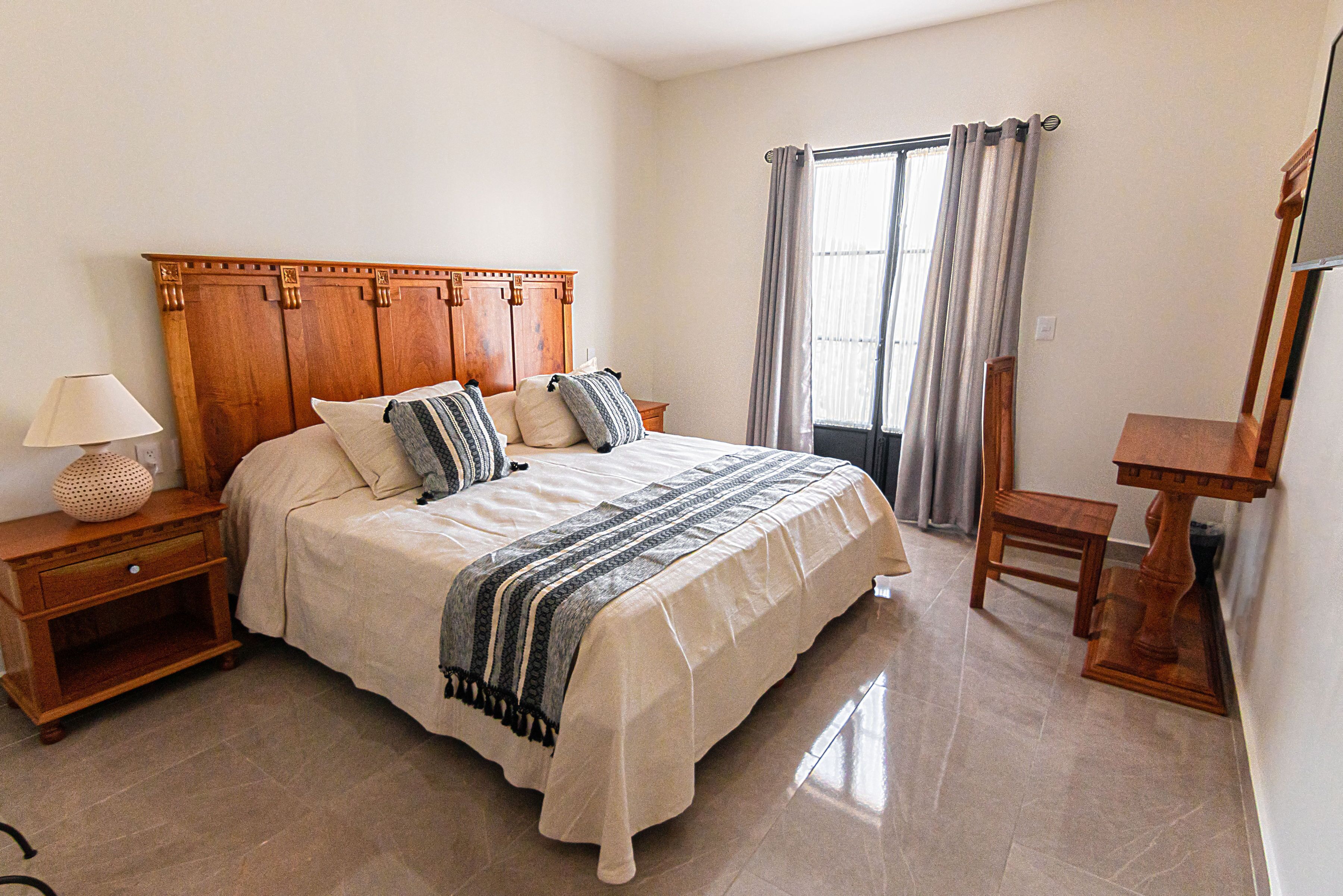 Un dormitorio con cabecera de madera, cama con manta a rayas, mesita de noche con lámpara, ventana con cortinas y una silla de madera.