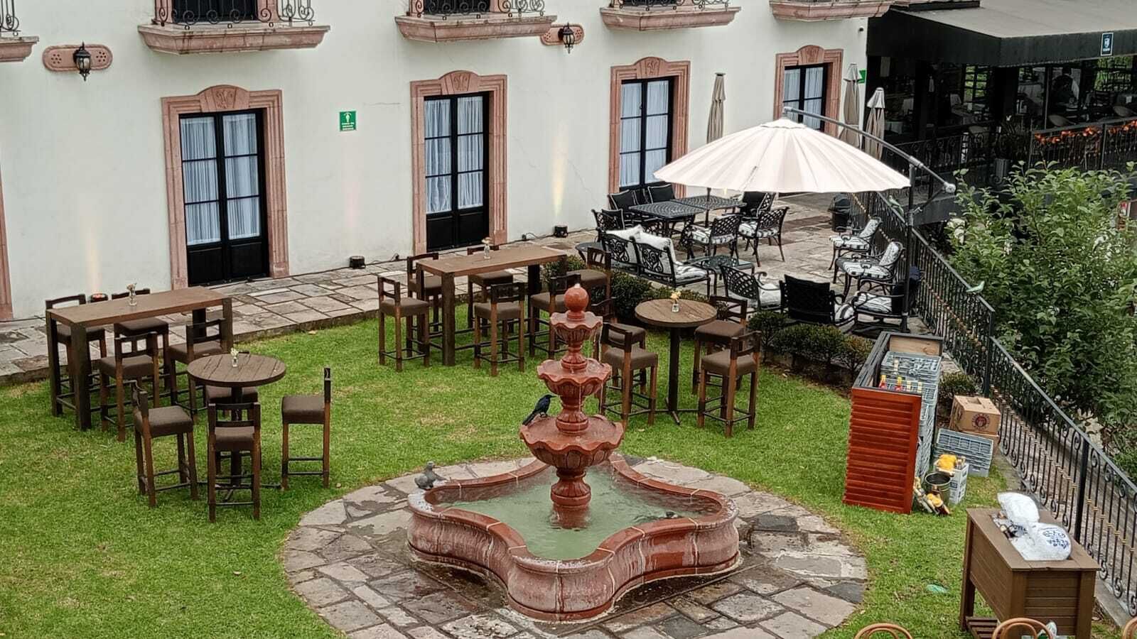 Eine Terrasse im Freien mit Tischen und Stühlen, einer zentralen Fontäne und einem Gebäude mit mehreren Fenstern und Balkonen.