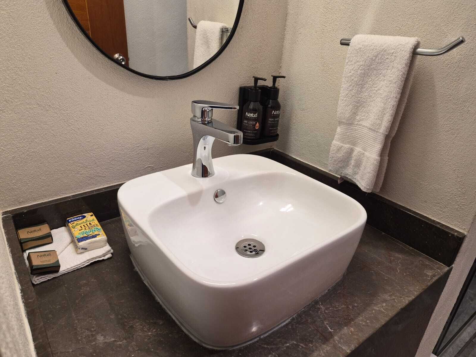 Un baño con un lavabo blanco, un espejo, un perchero para toallas y artículos de aseo.