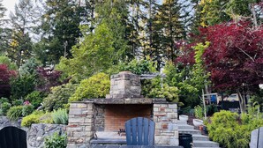 Chimenea de piedra al aire libre con un banco de madera y una silla frente a ella, rodeada de diversos árboles y arbustos.