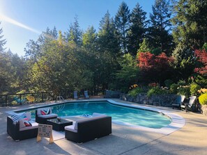 A swimming pool with lounge chairs and a small table, surrounded by trees and a stone wall. - Magical Farm Villa with Heated Salt Water Pool (Duncan)