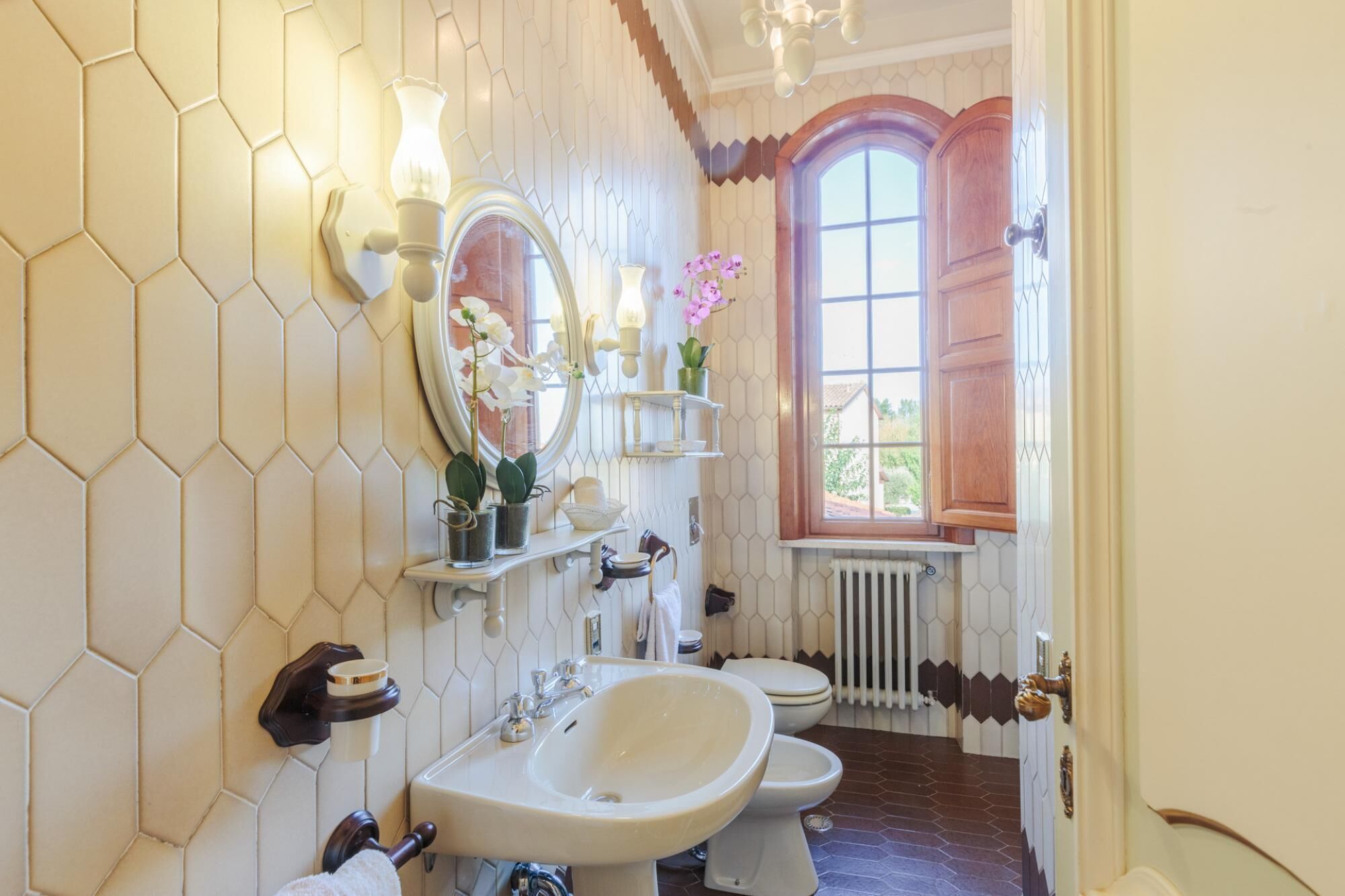 A bathroom with a hexagonal tile wall, a large mirror, a sink, a toilet, and a window with wooden shutters.
