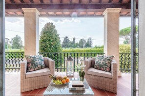 A covered patio with wicker furniture, a glass table with fruit and books, and a view of greenery and trees. - Villa Cesare Convenient and Smart (Lucca)