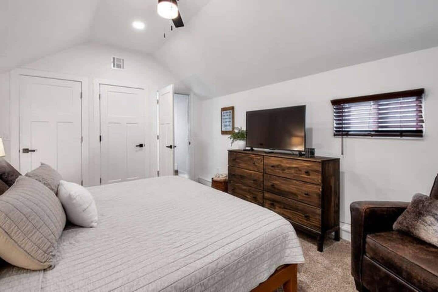 Une chambre à coucher avec un grand lit, une commode en bois, un téléviseur et une fenêtre avec des stores.