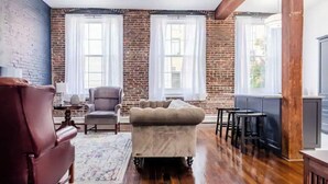 A living room with a brick wall, two windows with white curtains, a leather armchair, a sofa, and a wooden floor. - Downtown close to UAB, St. Vincent’s, BJCC and Airport  (Birmingham)