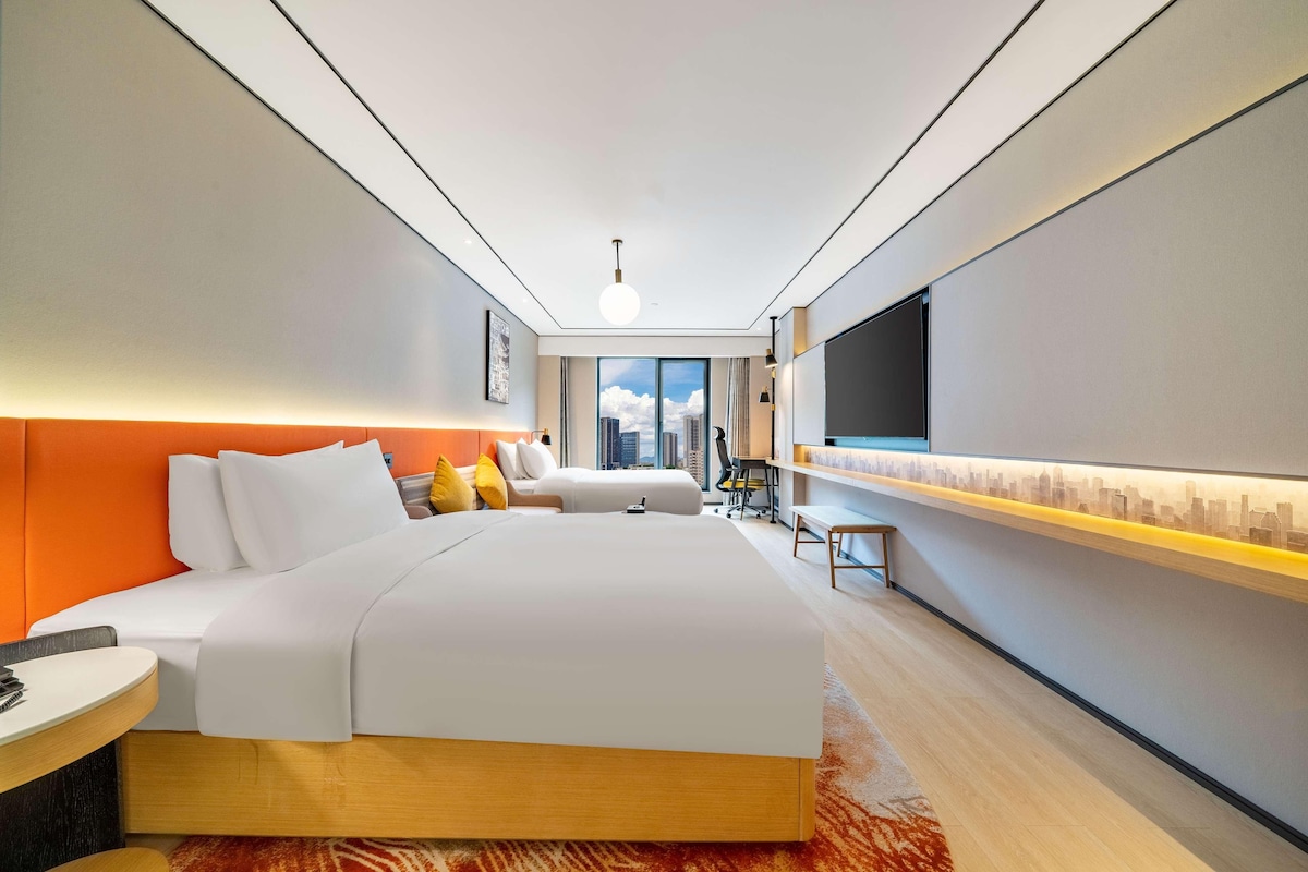 A modern hotel room with a large bed, a desk, a chair, a TV, and a city view.