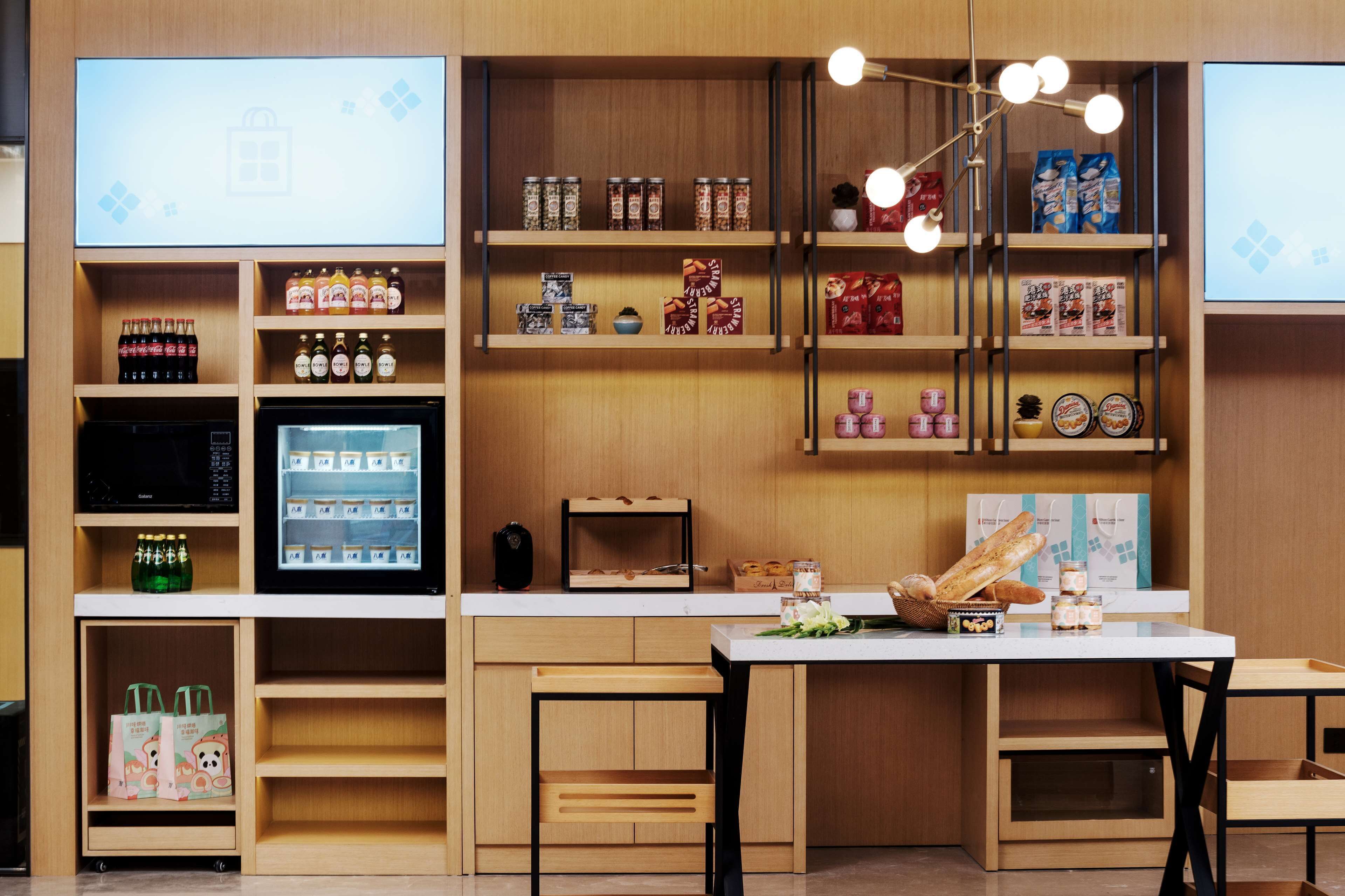 A kitchenette with a refrigerator, shelves with food items, a table with bread, and a coffee machine.