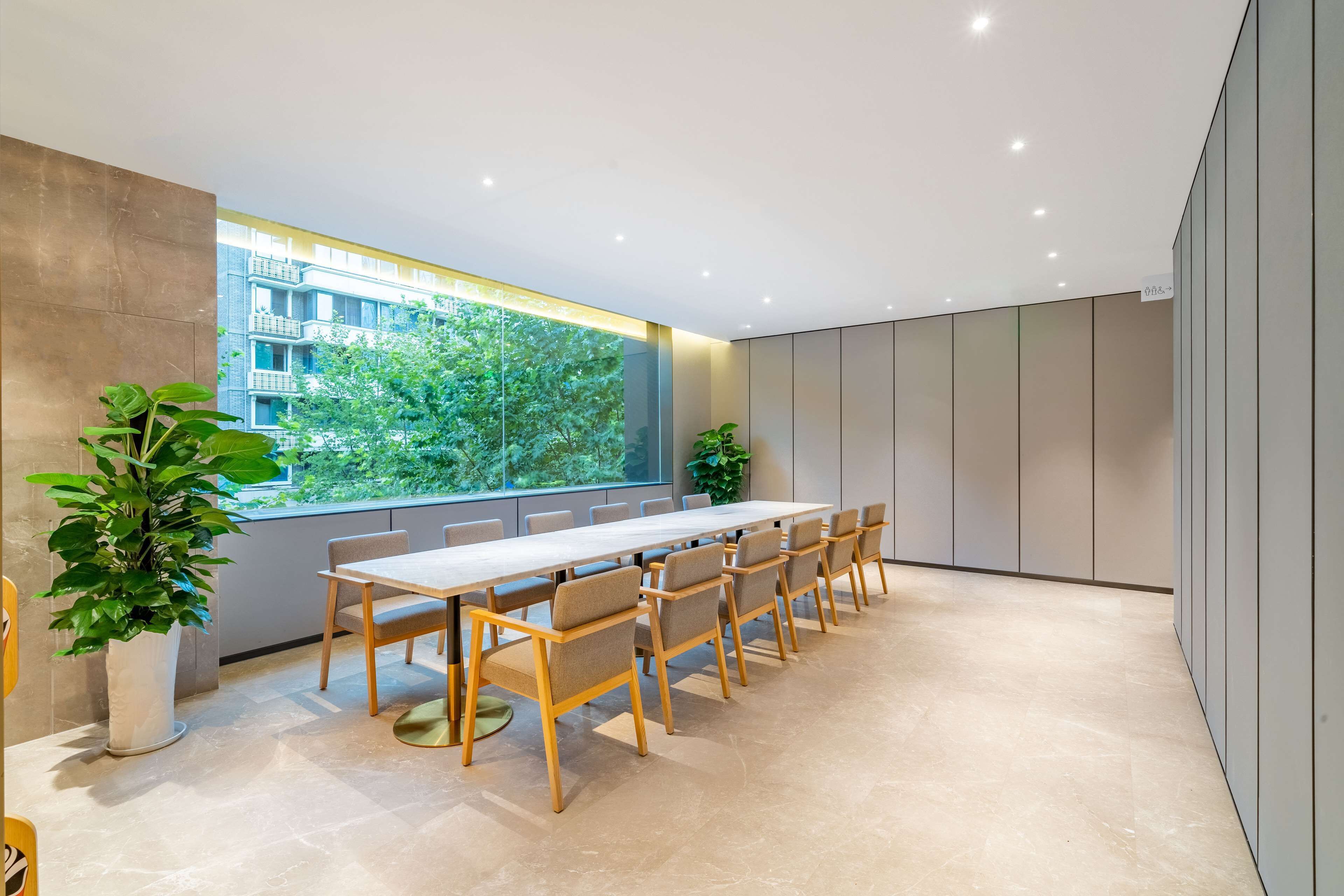 Una sala de reuniones moderna con una larga mesa y sillas, ventanas amplias con vistas a zonas verdes y edificios, y un diseño limpio y minimalista.