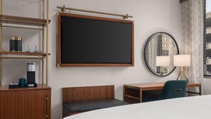 A hotel room with a bed, a desk, a chair, a bedside table with a lamp, a mirror, and a shelf with a coffee maker and glasses.