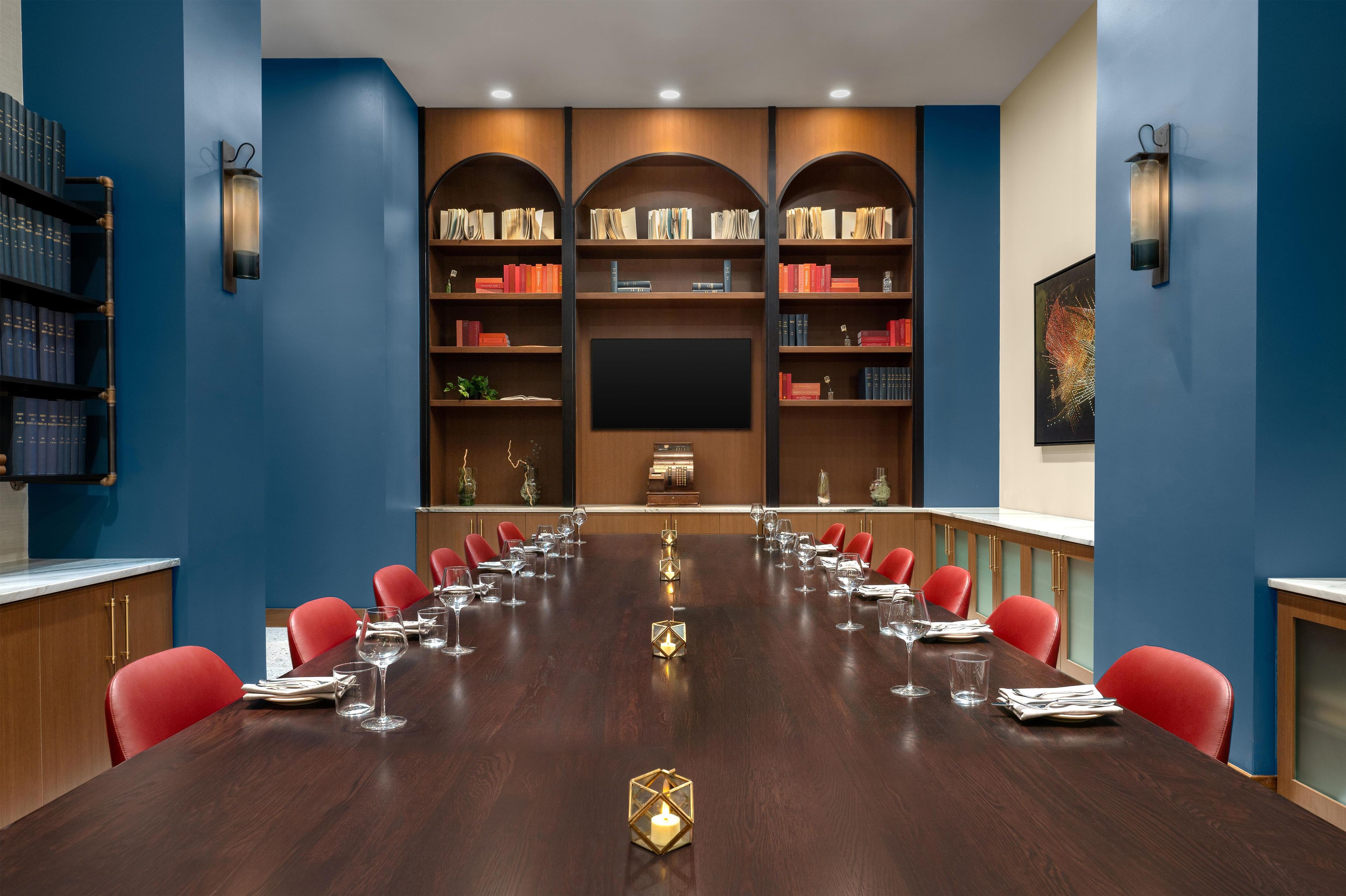 Un salón de conferencias con una larga mesa, sillas rojas y una estantería con libros y objetos decorativos.