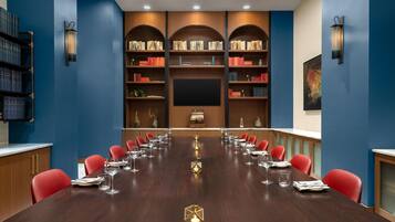 Un salón de conferencias con una larga mesa, sillas rojas y una estantería con libros y objetos decorativos.