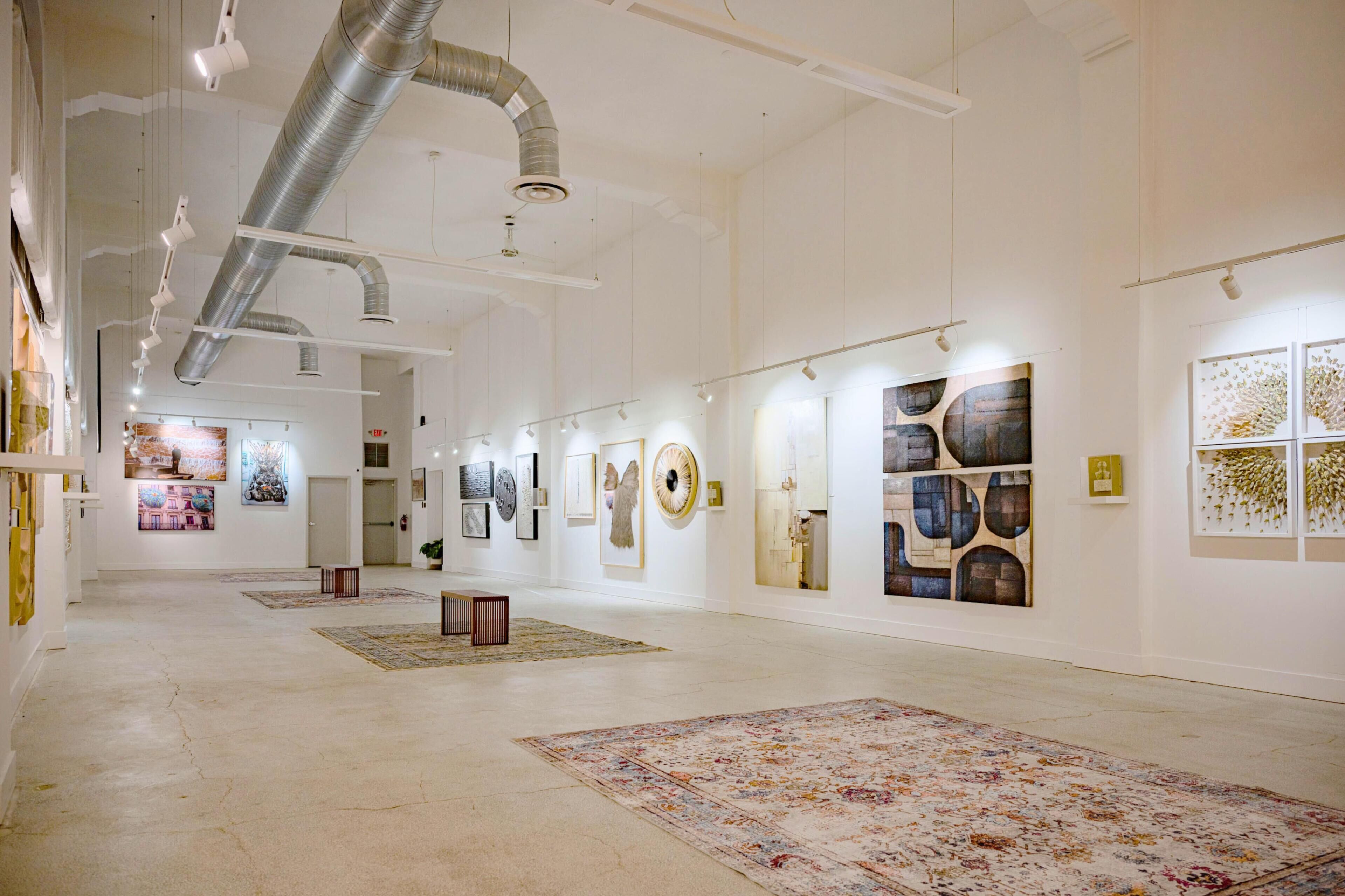 Una galería de arte con paredes blancas, conductos en el techo y obras expuestas en las paredes.