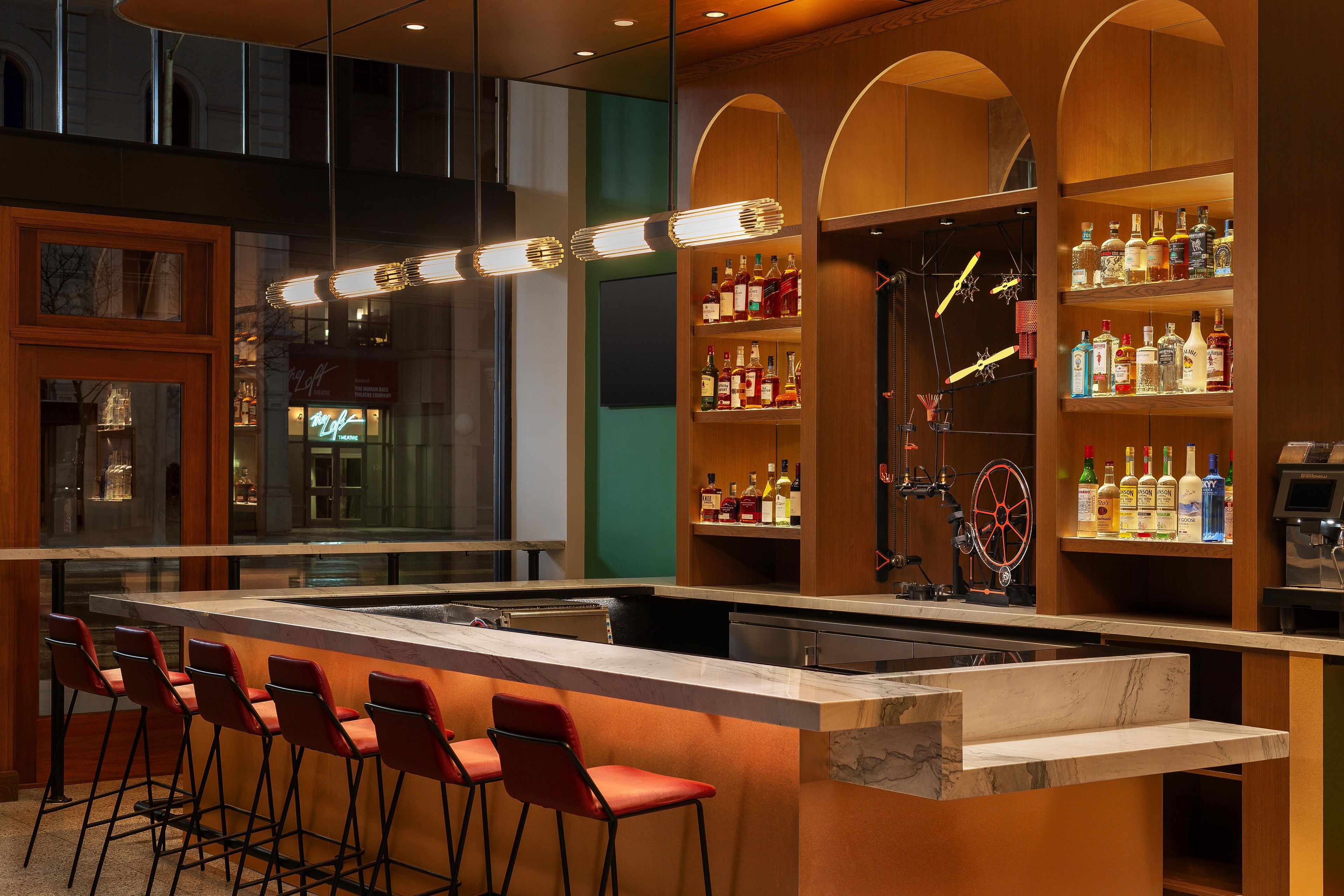 Zona de bar con una barra alargada, taburetes rojos y estantes llenos de botellas.