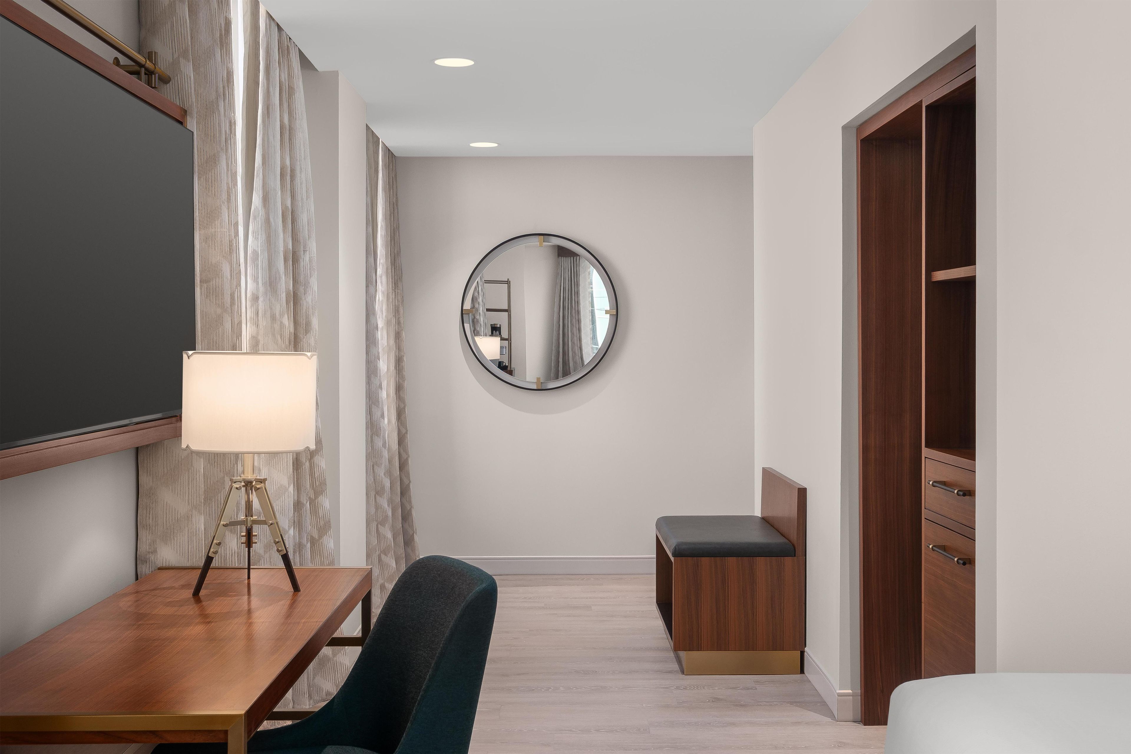 A hotel room with a wooden desk, a chair, a bedside table with a lamp, a round mirror, and a wardrobe.