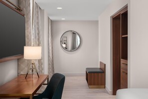 A hotel room with a wooden desk, a chair, a bedside table with a lamp, a round mirror, and a wardrobe. - Hotel Ardent Dayton Downtown, Tapestry Collection By Hilton (Dayton)