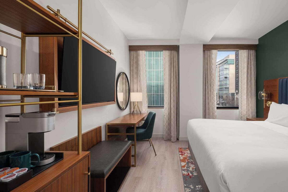 Ett hotellrum med en säng, ett skrivbord med en kaffemaskin, en stol, en tv och ett fönster med gardiner.