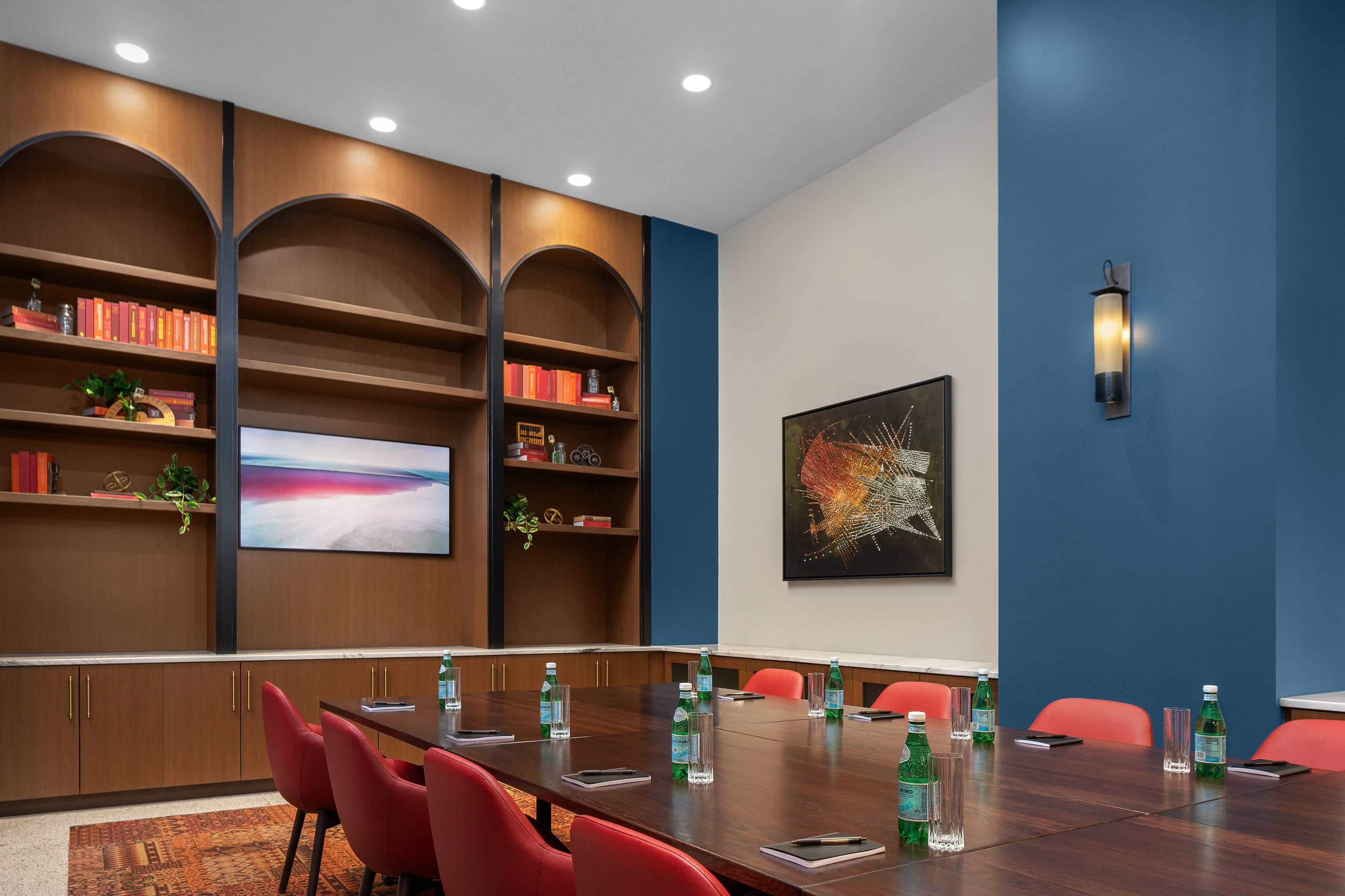 Un salón de conferencias con una mesa larga, sillas rojas y una estantería con libros y objetos decorativos.