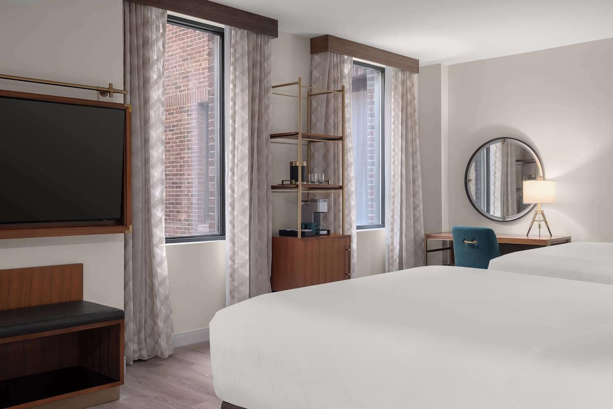 Ett hotellrum med en stor säng, ett skrivbord med en stol, en bokhylla, en spegel och ett fönster med gardiner.