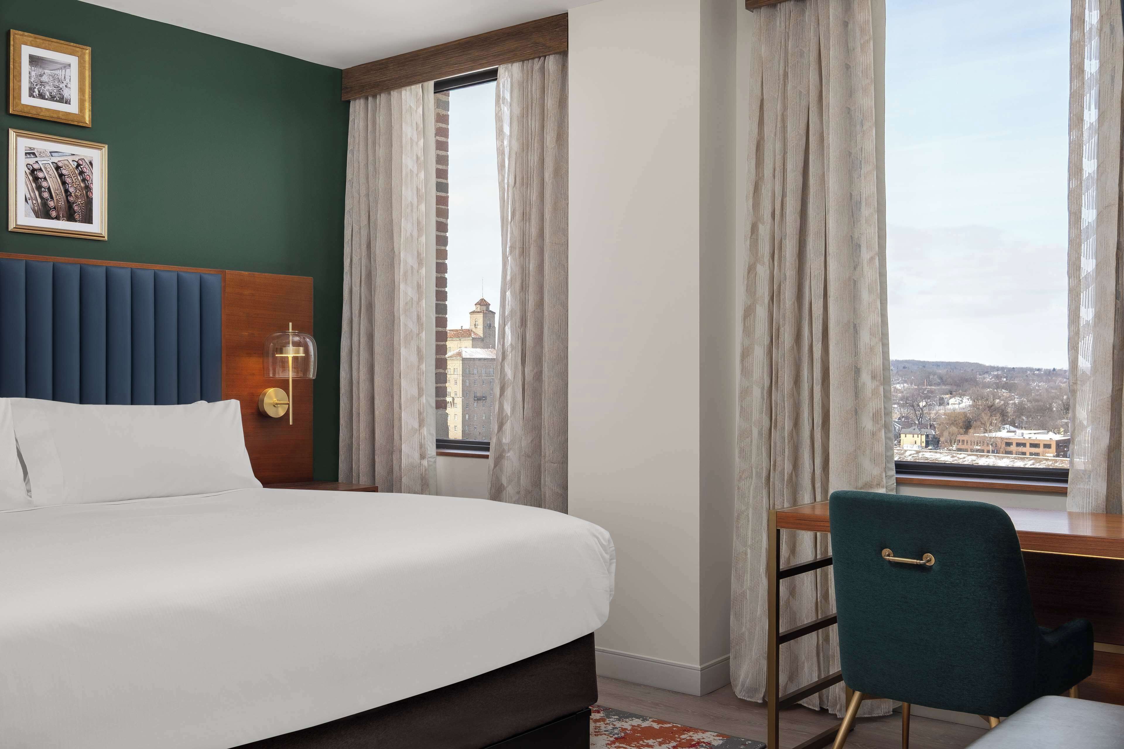Una camera d'albergo con un letto, una scrivania e una sedia. Alle finestre sono presenti delle tende e un quadro appeso al muro.