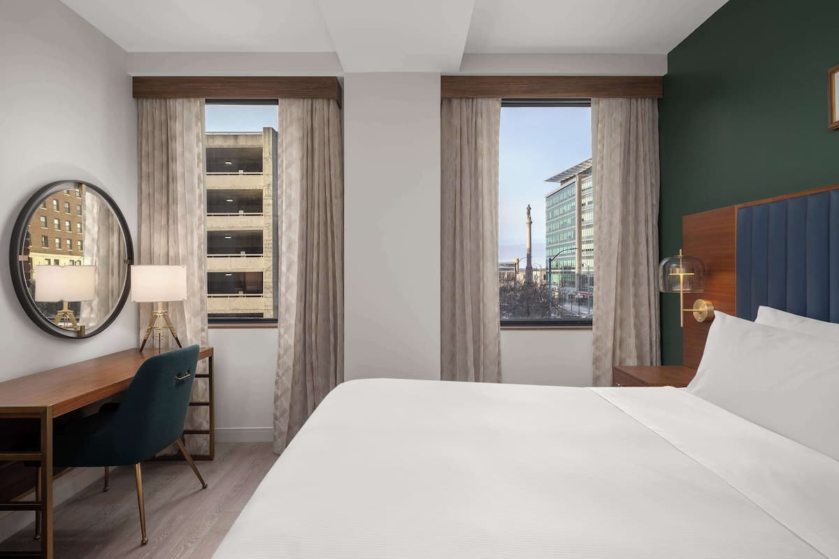 Ett hotellrum med en säng, ett skrivbord, en stol, två fönster med gardiner, en rund spegel och utsikt över byggnaderna utanför.
