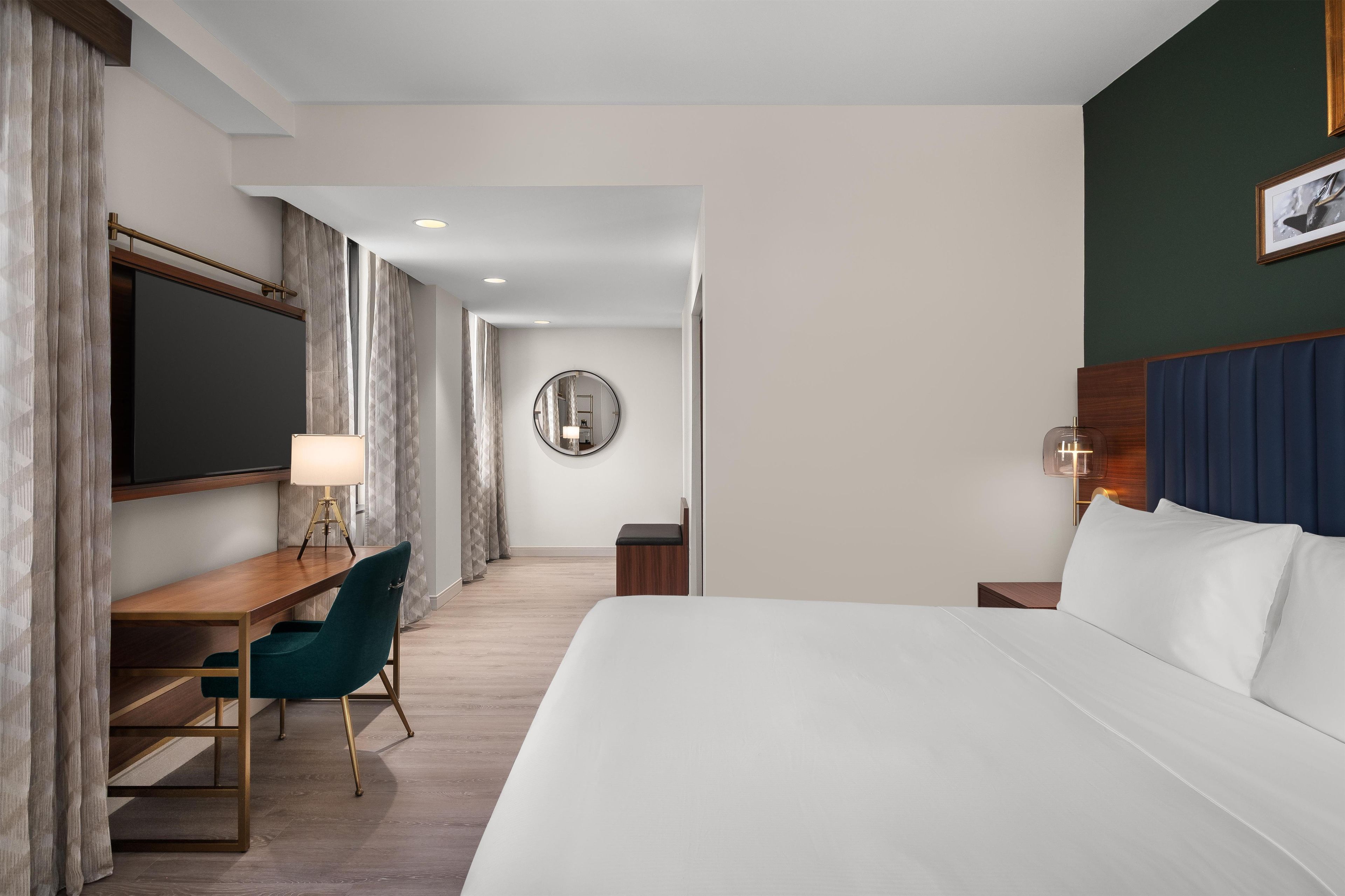 A hotel room with a bed, desk, chair, and a round mirror on the wall.