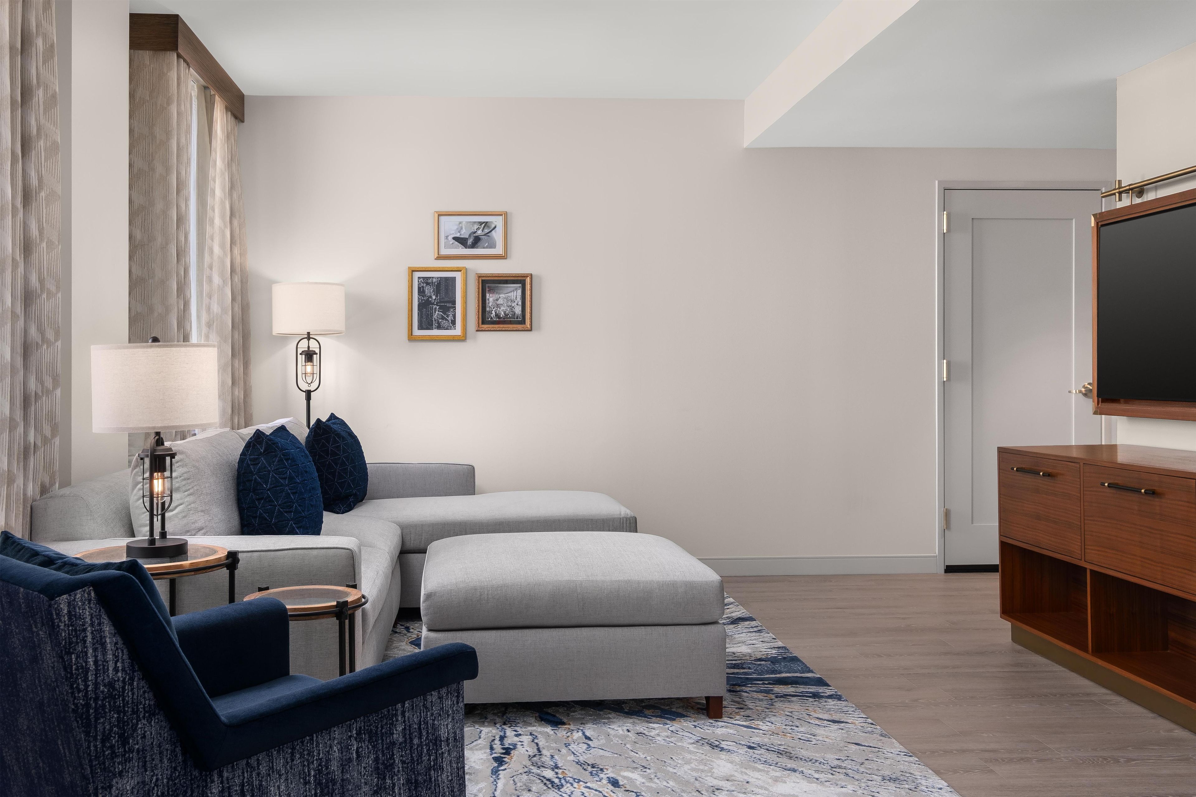 Een moderne woonkamer met een bank, een poef en een televisie. Er hangen ingelijste foto's aan de muur en een lamp op een bijzettafeltje.