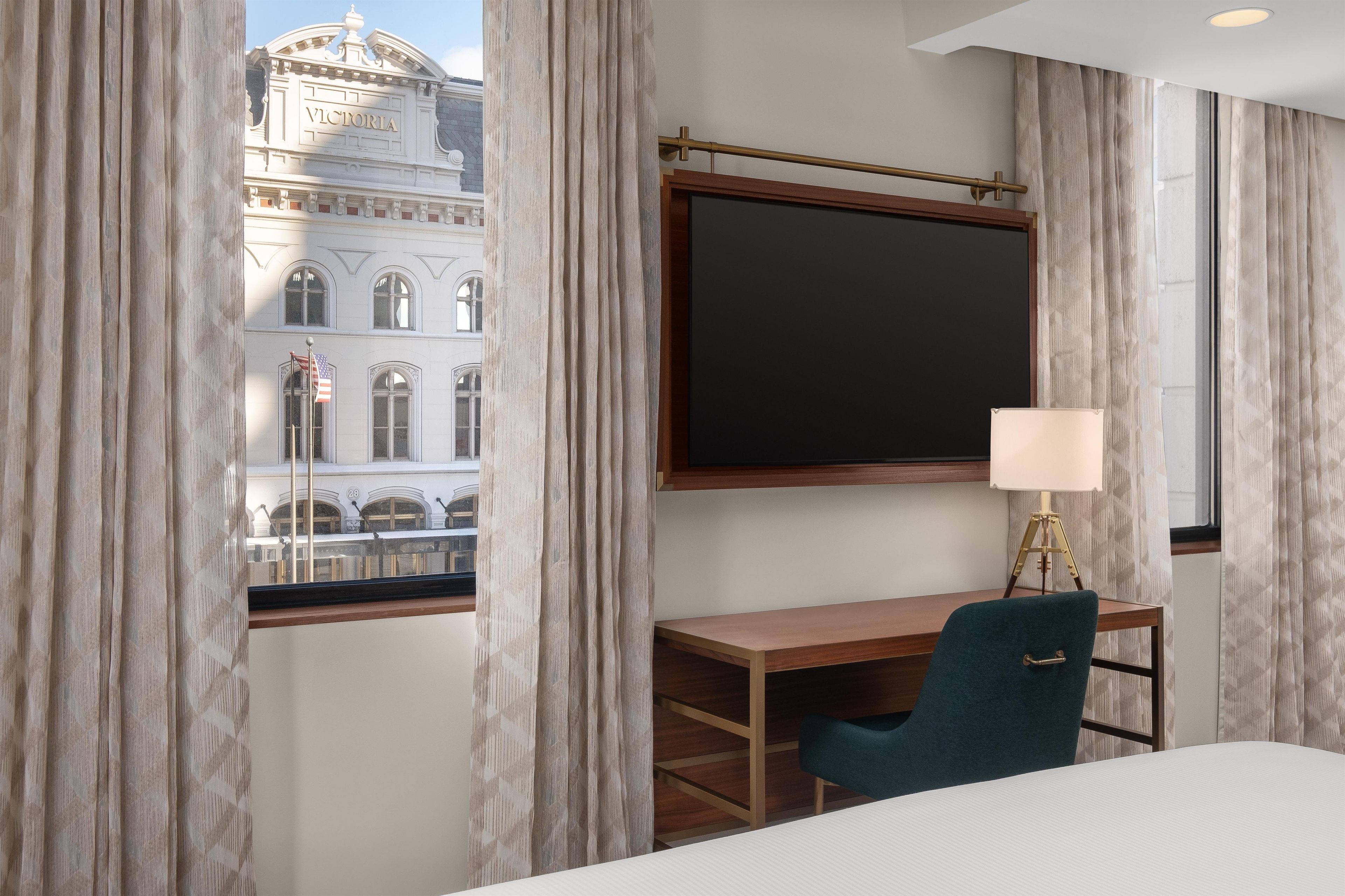 A hotel room with a large window, a desk, a chair, and a bed.